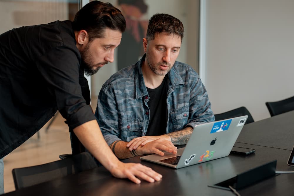 Dos hombres colaboran en una laptop plateada. Uno está sentado y el otro está de pie, inclinado sobre la mesa, observando la pantalla.