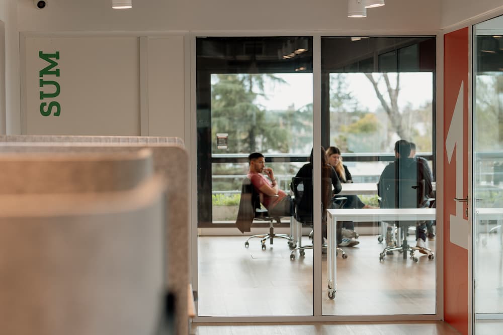 Una vista a través de una pared de cristal hacia una sala de reuniones donde se ven personas sentadas alrededor de una mesa. En primer plano, se ve parte de un letrero verde con la palabra 'SUM' en una pared.