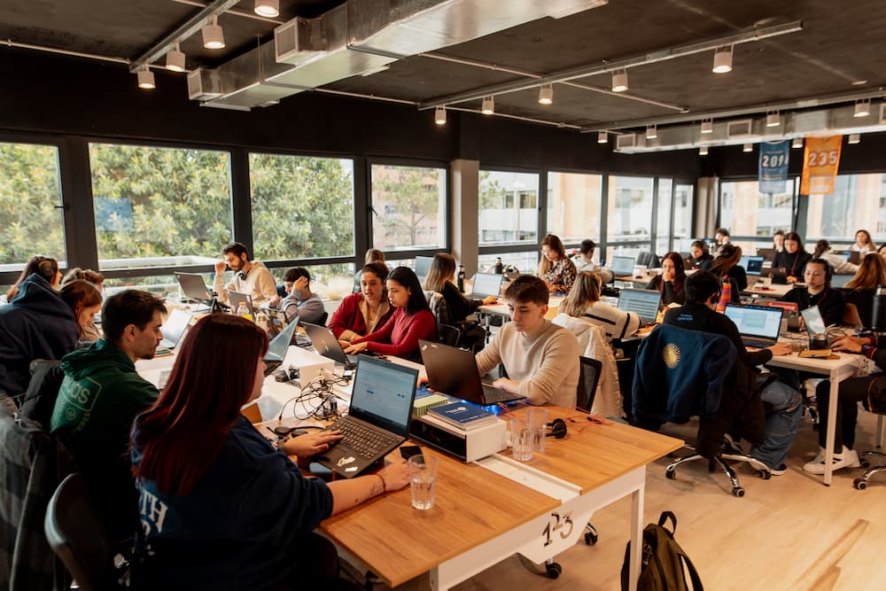 Una oficina grande y moderna con muchas personas trabajando en sus laptops en escritorios de madera. Grandes ventanas ofrecen vistas al exterior.