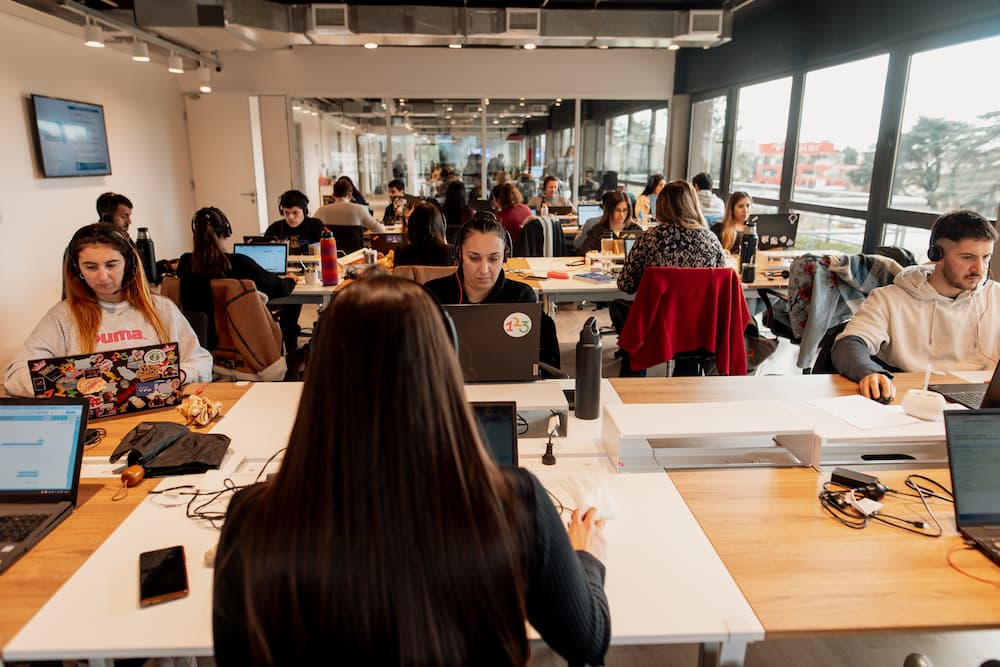 Una vista desde detrás de una mujer en una oficina abierta, mostrando a varias personas trabajando en sus laptops. El espacio es amplio con grandes ventanas en el fondo.