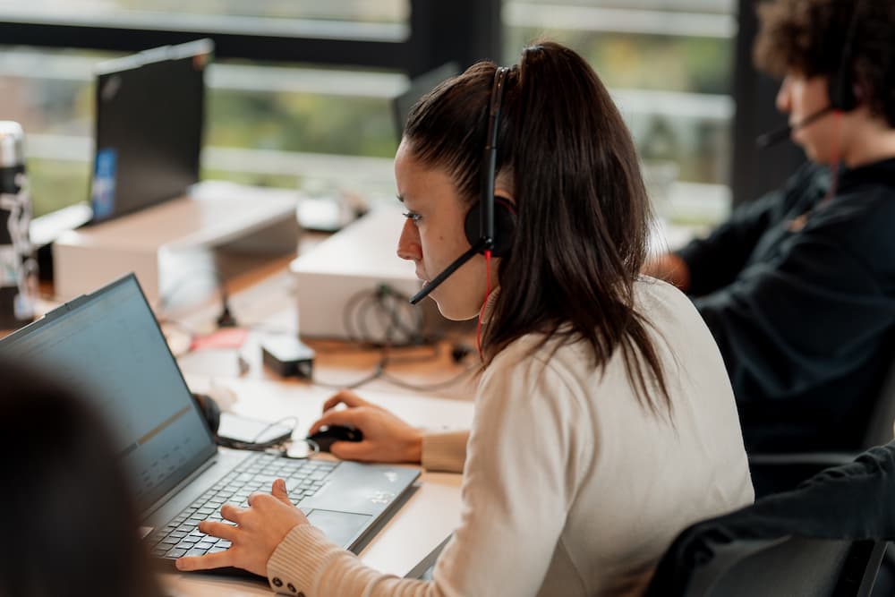 Una mujer con auriculares y micrófono trabaja en su laptop en una oficina. Está concentrada en la pantalla, con su mano en el ratón y la otra en el teclado.