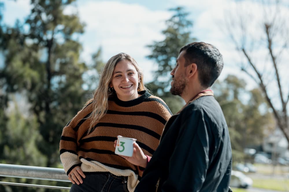 Un hombre y una mujer sonríen mientras conversan al aire libre, posiblemente en una terraza. La mujer lleva un suéter a rayas y el hombre sostiene una taza con el número '3'. Hay árboles y cielo azul de fondo.