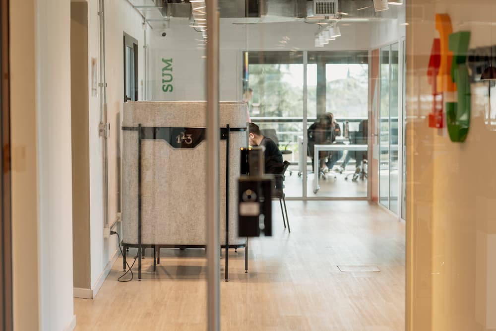 Una vista a través de una pared de cristal en una oficina moderna. Se ve un espacio de trabajo con una persona sentada al fondo, y en primer plano, divisores de oficina con un letrero que dice 'SUM' en la pared.