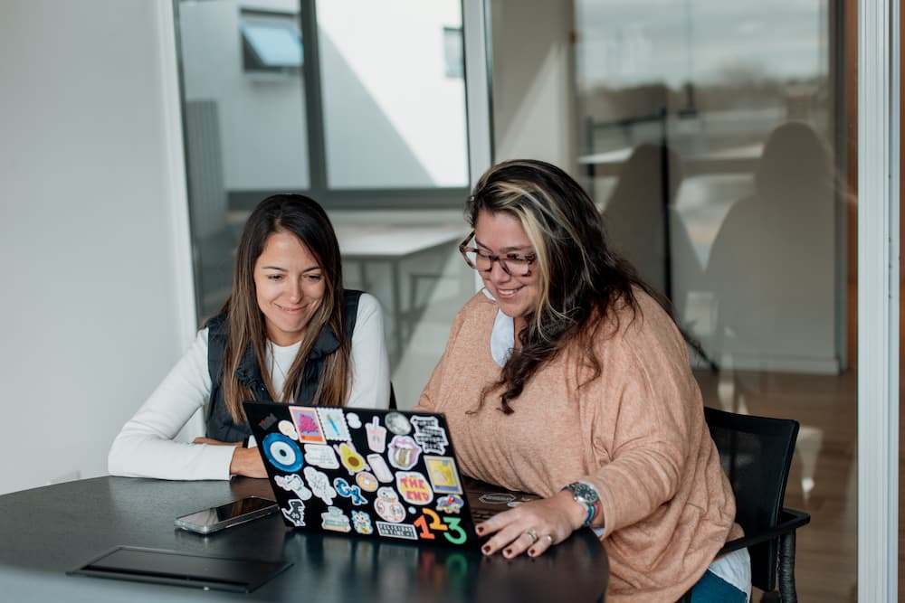 Dos mujeres miran una laptop llena de pegatinas en una oficina. La mujer de la derecha, que lleva gafas, sonríe y señala la pantalla.