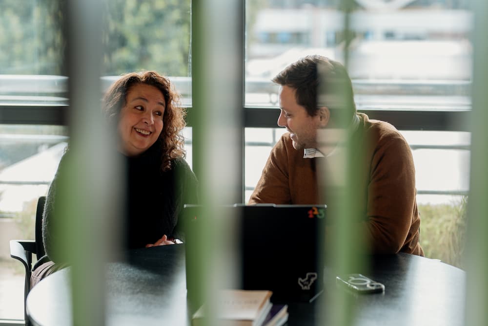 Un hombre y una mujer sonríen mientras conversan, sentados frente a una laptop. La imagen está ligeramente desenfocada por elementos en primer plano.