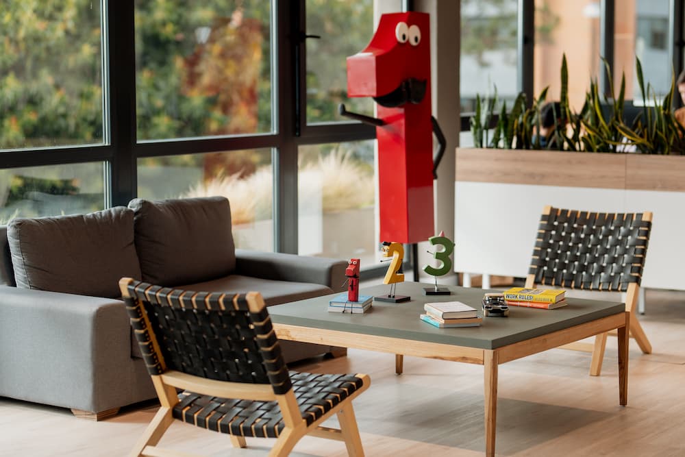 Un rincón de oficina con un sofá gris y dos sillas con respaldo de mimbre. Una mesa de centro tiene figuras decorativas de los números 1, 2 y 3, junto con libros. Plantas en macetas se ven al fondo cerca de una ventana.
