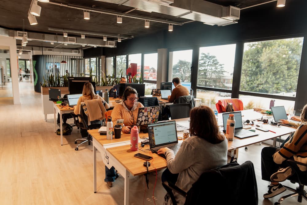 Un plano general de una oficina moderna y luminosa con varias personas trabajando en escritorios individuales con laptops. Grandes ventanas ofrecen vistas al exterior.