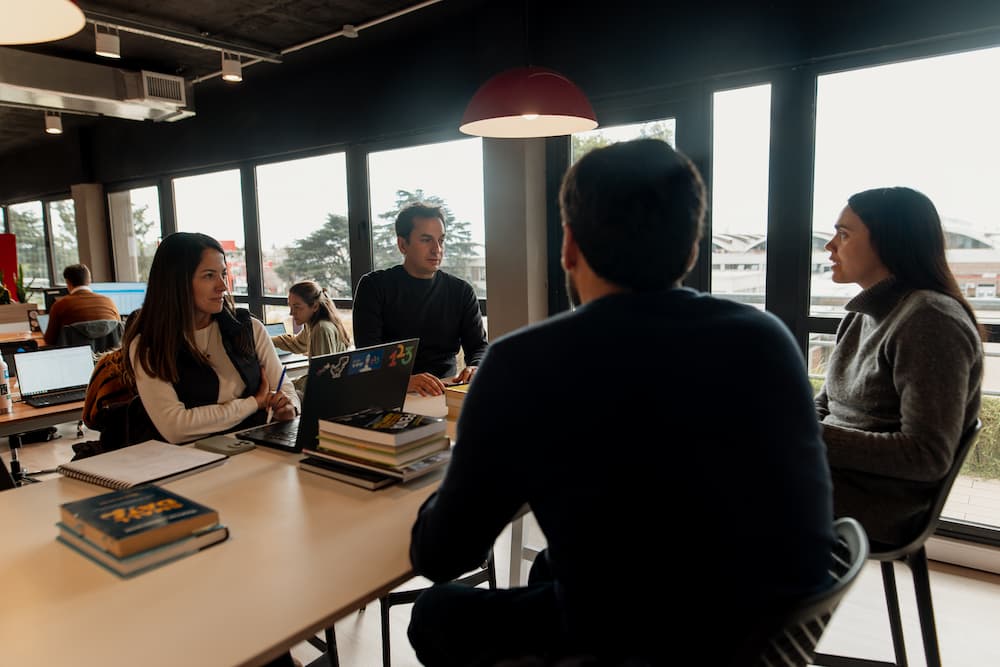 Un grupo de cuatro personas conversa alrededor de una mesa en una oficina abierta. Hay laptops y libros sobre la mesa, y grandes ventanas en el fondo.